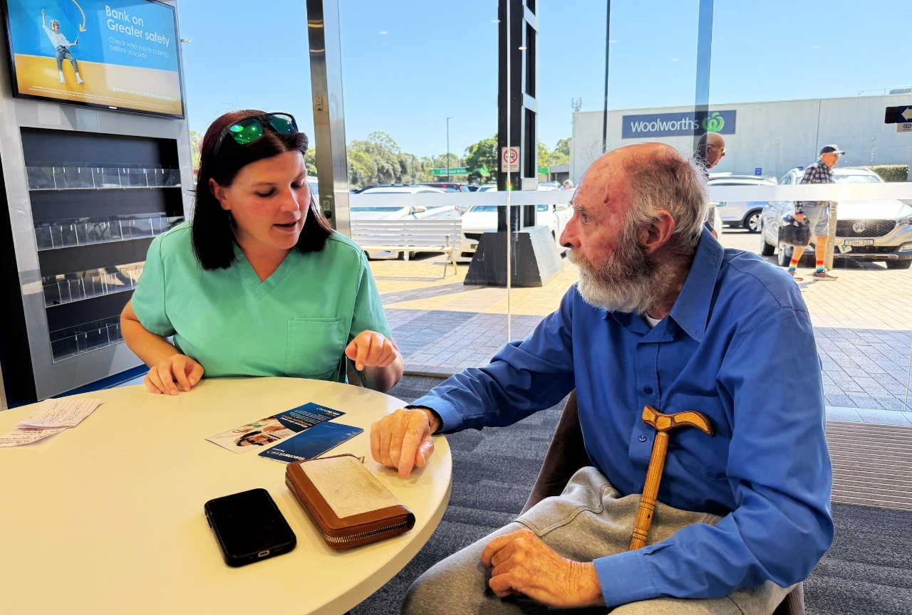 Kiri sitting with John Bell at a table outside a bank, helping with his banking needs