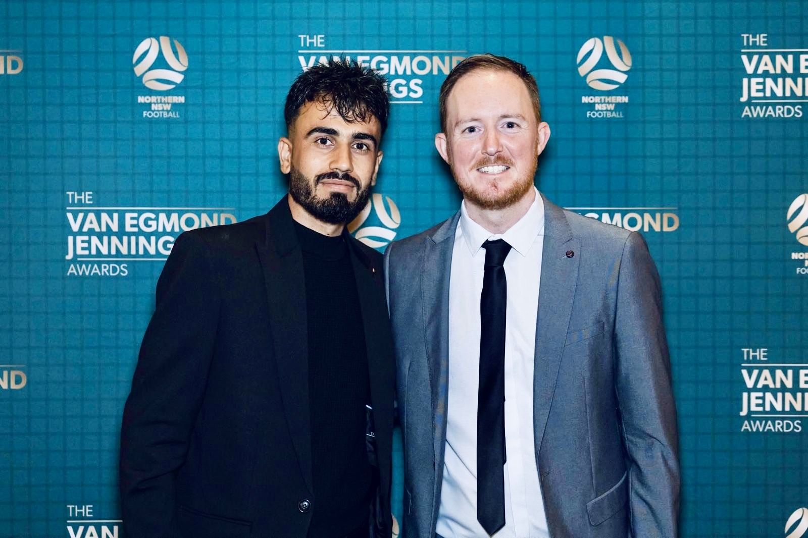 Mustafa in a black suit standing with Joseph Wright at the Van Egmond Jennings Awards, teal backdrop
