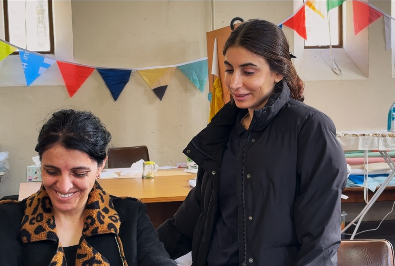 Orien standing over a laughing seated woman at the sewing table in Mosaic's women's group, Newcastle